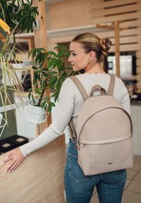Beige leather backpack with a rounded shape, thin straps, and a front zipper pocket. Textured finish and a small logo at the base.