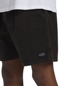 Bermudas de algodón negras con una superficie texturizada, dobladillo cosido y un parche de marca morado en el lado izquierdo.