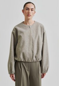 Beige zip-up jakke med rund hals, elastisk i bunnen og ballongsleeves, laget av et mykt, teksturert materiale. Båret over grønne bukser.