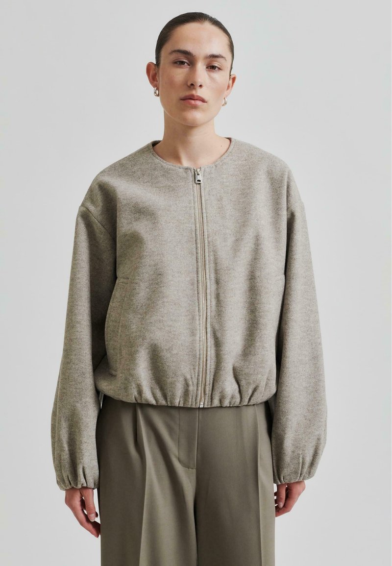 Beige zip-up jakke med rund hals, elastisk i bunnen og ballongsleeves, laget av et mykt, teksturert materiale. Båret over grønne bukser.