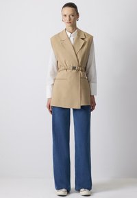 Beige mouwloze vest met een ingezette taille, gelaagd over een wit shirt, gecombineerd met wijduitlopende blauwe denim jeans en witte sneakers.