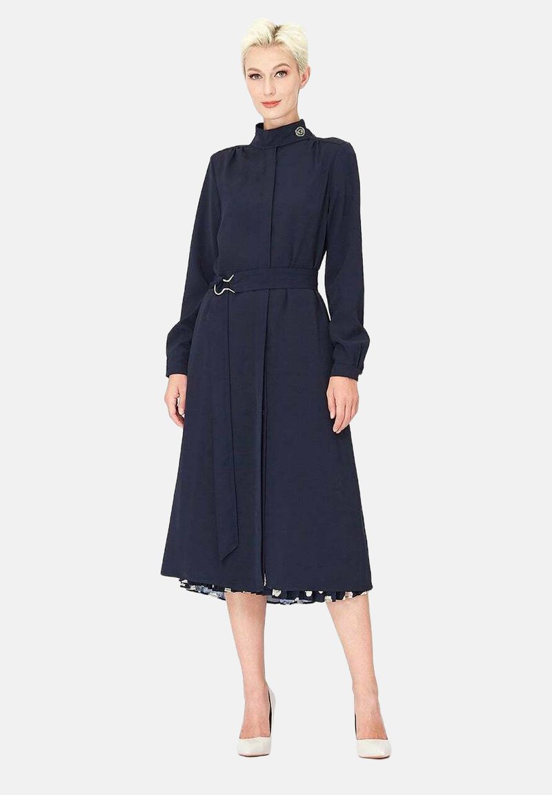 Robe bleu marine à manches longues, col montant, taille ceinturée et jupe évasée. Présente un bouton décoratif et une doublure à motifs contrastants.