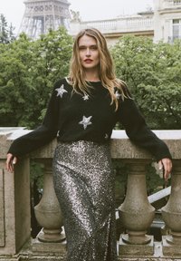 Juoda megzta palaidinė su sidabriniais žvaigždžių akcentais, derinama su blizgančia sidabrine sijonu. Modelis stovi prie akmeninės turėklos, o fone matyti žaluma.