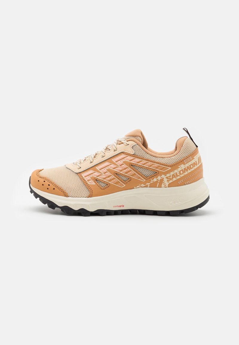 Chaussure de trail beige et marron clair avec une tige en mesh, des surpiqûres synthétiques en zigzag, une semelle épaisse avec crampons noirs et une languette à l’arrière.