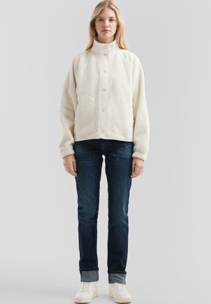 Jeune femme portant une veste blanche à boutons texturée, un jean bleu foncé avec les poignets retroussés et des baskets blanches, se tenant devant un fond clair uni.