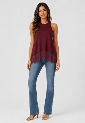 Donna che indossa una canotta bordeaux senza maniche con orlo in pizzo, jeans blu e tacchi beige, in piedi davanti a uno sfondo grigio chiaro semplice.