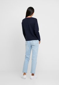 Pull marin avec détail de col rouge, associé à un jean bleu clair et des baskets blanches. La tenue présente des manches longues et une coupe décontractée.