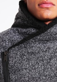 Abrigo gris de mezcla de lana con un cuello alto y detalles negros. Presenta una superficie texturizada y un cierre de cremallera lateral.