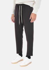 Uomo in pantaloni neri a tubo con cordino, sneakers bianche con suole beige, calzini neri e un maglione bianco a coste, in piedi su uno sfondo bianco.