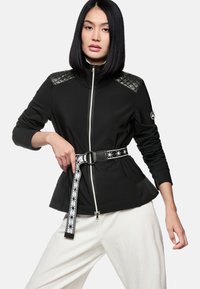 Schwarze Sportjacke mit gesteppten Schulterpartien, silbernem Reißverschluss und einer taillierten Taille, die durch einen gemusterten Gürtel betont wird; getragen über weißen Hosen.