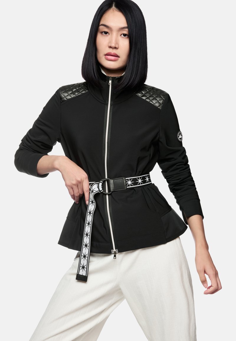 Schwarze Sportjacke mit gesteppten Schulterpartien, silbernem Reißverschluss und einer taillierten Taille, die durch einen gemusterten Gürtel betont wird; getragen über weißen Hosen.