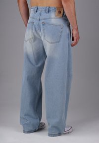 Hellblaue weit geschnittene Jeans mit einem ausgewaschenen Finish, featuring hinteren Taschen und einem braunen Lederlogo-Aufnäher am Bund. Weiße Sneakers sichtbar.