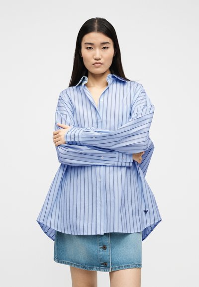 Νεαρή γυναίκα με μακριά σκουρόχρωμα μαλλιά φοράει μια φαρδιά μπλε ριγέ πουκάμισο με κουμπιά και μια φούστα από τζιν, στεκόμενη με σταυρωμένα τα χέρια.