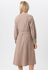 Beige midi-jurk met lange mouwen, voorzien van oogjeskant details op de schouders en manchetten, en een stoffen riem om de taille.