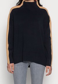 Pull en maille bleu marine à col montant, avec des rayures texturées dorées le long des manches. Coupe ample avec ourlet et poignets côtelés.