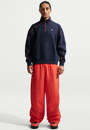 Mann steht und trägt ein marineblaues Sweatshirt mit Viertel-Reißverschluss und roter Paspel, leuchtend rote, weit geschnittene Hose und rot-schwarze Sneakers vor weißem Hintergrund.