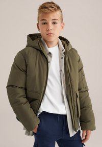 Puhastna jakna v olivno zeleni barvi, z kapuco, zadrgo in stranskimi žepi. Nosimo čez belo srajco in sivo pločevinasto srajco, z mornarsko modrimi hlačami.