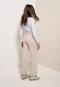 Nainen, jolla on pitkät hiukset, pukeutuneena valkoiseen pitkähiharaiseen paitaan, beessiin leveälahkeisiin housuihin ja valkoisiin lenkkareihin, seisoo huoneen nurkkaa kohti neutraalin sävyisessä huoneessa.