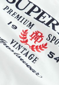 Bílá látka s námořnickými a červenými vyšívanými nápisy. Designy zahrnují "SUPER" a "VINTAGE" s červeným vavřínovým grafikou.
