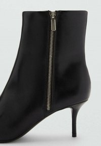 Bottine en cuir noir avec un talon haut élancé, dotée d'une fermeture éclair latérale en argent et d'une texture lisse. Design simple et élégant sans embellissements supplémentaires.