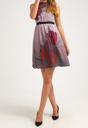 Mujer con vestido púrpura sin mangas con un gran estampado floral rojo, cinturilla negra y tacones altos negros, de pie con las piernas cruzadas.