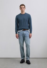 Hombre vestido con un suéter azul de manga larga, jeans azul claro con un cinturón marrón y zapatos negros, de pie contra un fondo sencillo.