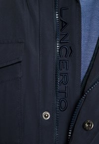 Marynarka w kolorze granatowym z zapięciem na zamek błyskawiczny, widocznym haftowanym logo, teksturowanym materiałem oraz detalami z zatrzaskami. Podszewka z wzorzystej tkaniny.