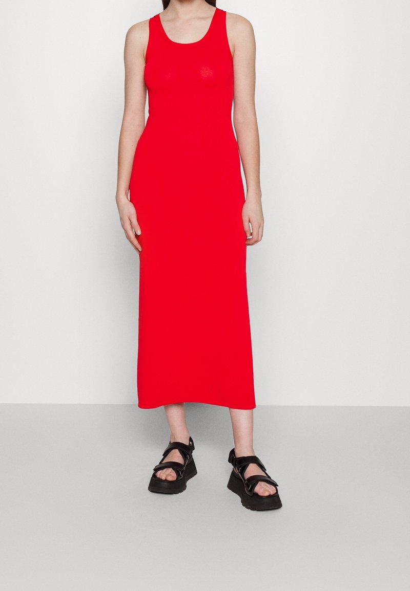 Robe longue rouge sans manches en tissu extensible, silhouette ajustée, couleur unie, associée à des sandales noires massives avec des brides.