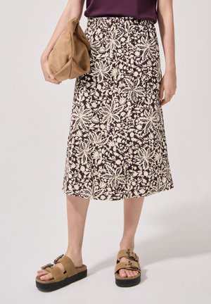 Kvinde iført en brun og cremefarvet blomsterprintet midi-nederdel, tanfarvede platformsandaler med spænder og holder en tanfarvet blød clutchtaske.