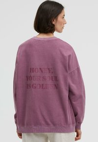 Überdimensioniertes, lila Sweatshirt aus weichem Material. Mit erhabenem Text auf der Rückseite: "HONEY, DEINE SEELE IST GOLDEN." Ripp-Ausschnitt und -Ärmel.