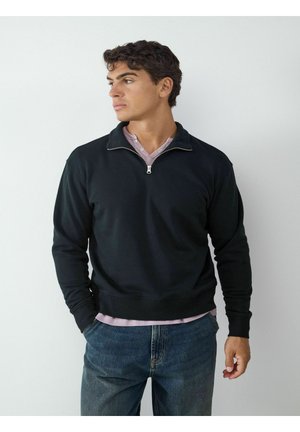 Junger Mann, der einen schwarzen Pullover mit Viertelreißverschluss über einem hellrosa Hemd und blauen Jeans trägt, schaut nach links, die Hände teilweise in den Taschen.