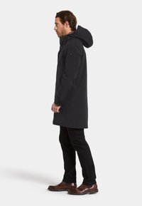 Svart vattentät rock med huva, midjehöjd, med knäppning fram och sydda ärmar. Bärs med svarta byxor och bruna skor.