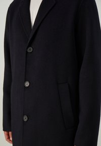 Man wearing a dark navy wool coat with three visible buttons over a light-colored ribbed sweater, standing against a plain background.