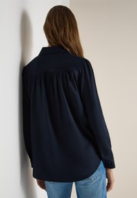 Femme aux longs cheveux bruns portant un chemisier ample à manches longues bleu marine foncé et un jean bleu, debout face à un mur clair.