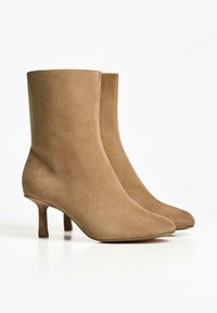 Botine din suede bej, cu vârf ascuțit și toc scurt, subțire. Designul are o textură fină și o siluetă minimalistă.