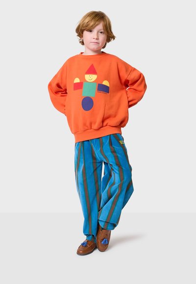 Narančasta dukserica s geometrijskim dizajnom lica, u kombinaciji s plavim prugastim hlačama s smešnim linijama. Smeđe cipele sa plavim resicama.