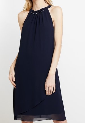 Mouwloze marineblauwe halterjurk met geplooide halslijn en kleine metalen kraaltjes, gedragen tot net boven de knieën.