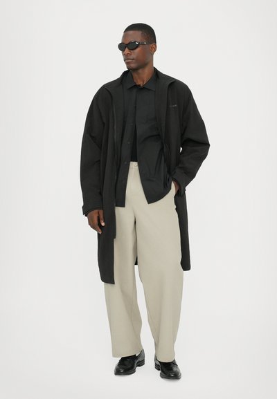 Black long coat over a black shirt, paired with beige trousers and black shoes. The figure wears dark sunglasses and stands on a plain background.