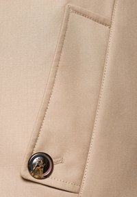 Beżowa tkanina o gładkiej fakturze z małą kieszonką naszywaną oraz okrągłym, metalowym guzikiem z detalem wzorniczym.