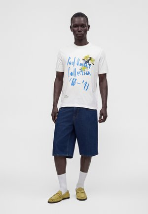 Man draagt een wit T-shirt met blauwe tekst en gele bloemen, donkerblauwe denim shorts, witte sokken en gele loafers, staand tegen een witte achtergrond.