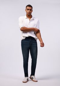 Camisa blanca de botones con mangas remangadas, combinada con pantalones vaqueros azul oscuro. Modelo lleva zapatillas blancas y negras, de pie contra un fondo neutro.
