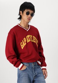 Red sweatshirt with "GAP ATHLETIC" in yellow, featuring a V-neck, white striped trim, paired with blue denim jeans and black sunglasses.