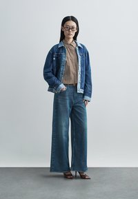 Denim jacket with light blue accents, striped brown and white shirt, loose blue jeans, and brown heeled mules against a neutral background.
