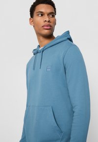 Blauwe hoodie van zacht stof, met een voorzak, trekkoord capuchon en een vierkante logopatch op de borst. Gladde textuur.