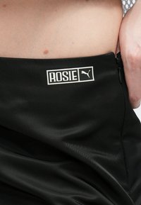 Crna atletska suknja s glatkom, sjajnom teksturom. Ima bijelu oznaku s logom "ROSIE" i Puma logom na struku. Uključena je detaljna zatvarač.