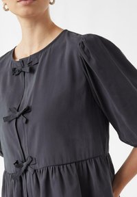 Dunkelgraues Kleid mit Puffärmeln, rundem Ausschnitt und zwei kleinen Schleifenakzenten vorne. Glattes, leichtes Material mit geraffter Taille.
