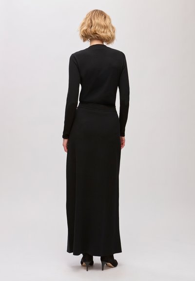 Femme aux cheveux courts et blonds, vêtue d'une longue robe noire et de chaussures à talons noirs, debout de dos sur un fond uni.