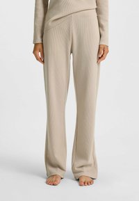 Persoon gekleed in beige, gestructureerde lange broek en bijpassend shirt, staande op blote voeten tegen een effen lichte achtergrond.