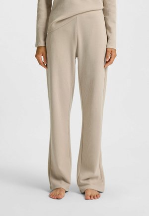 Person i beige strukturerade långbyxor och matchande topp, stående barfota mot en enkel ljus bakgrund.