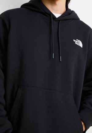 Person trägt schwarzen The North Face Hoodie mit Kordelzug und Fronttasche, zugeschnitten, um den Oberkörper und das Logo auf der Brust zu zeigen.